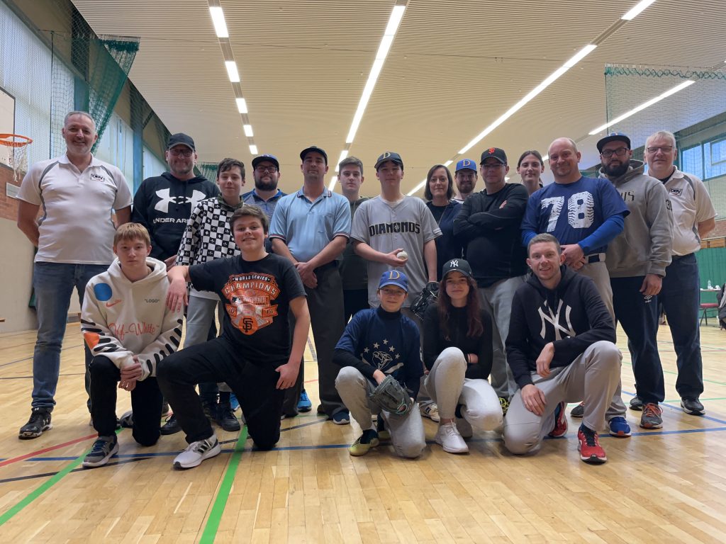 Eine Gruppe von Baseballspielern und Trainern posiert in einer Sporthalle für ein Gruppenfoto. Einige knien vorne, während andere hinten stehen.
