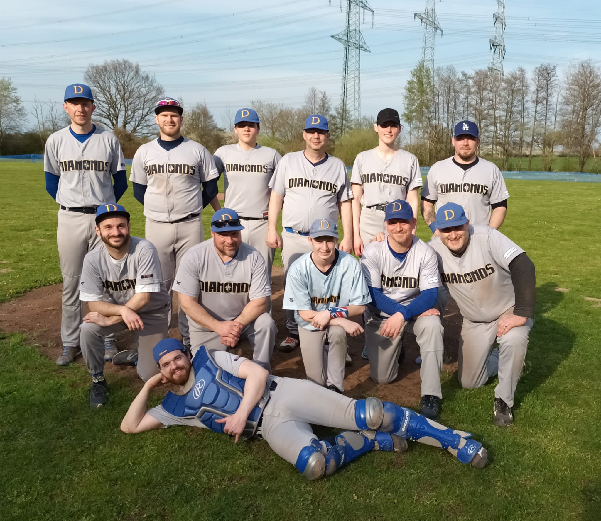 Eine Gruppe Baseballspieler in grauen Trikots steht und kniet für ein Teamfoto auf einem Baseballfeld, während einer von ihnen auf dem Boden liegt.