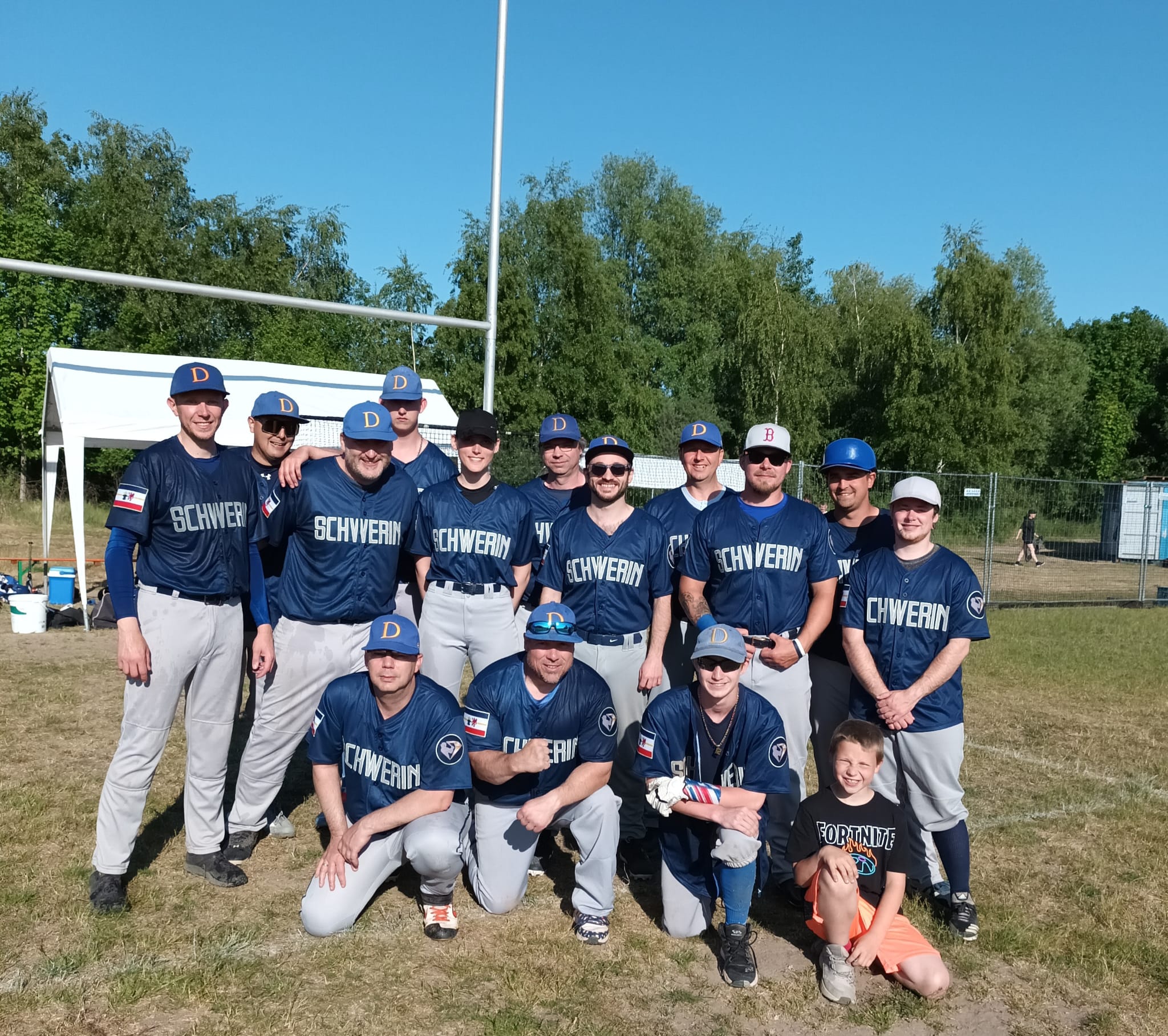Eine Gruppe von Baseballspielern in blauen Trikots und grauen Hosen steht auf einem Baseballfeld zusammen und posiert für ein Teamfoto. Vorne knien lächelnd drei Männer und ein Junge, während die Spieler hinter ihnen ebenfalls lachen.