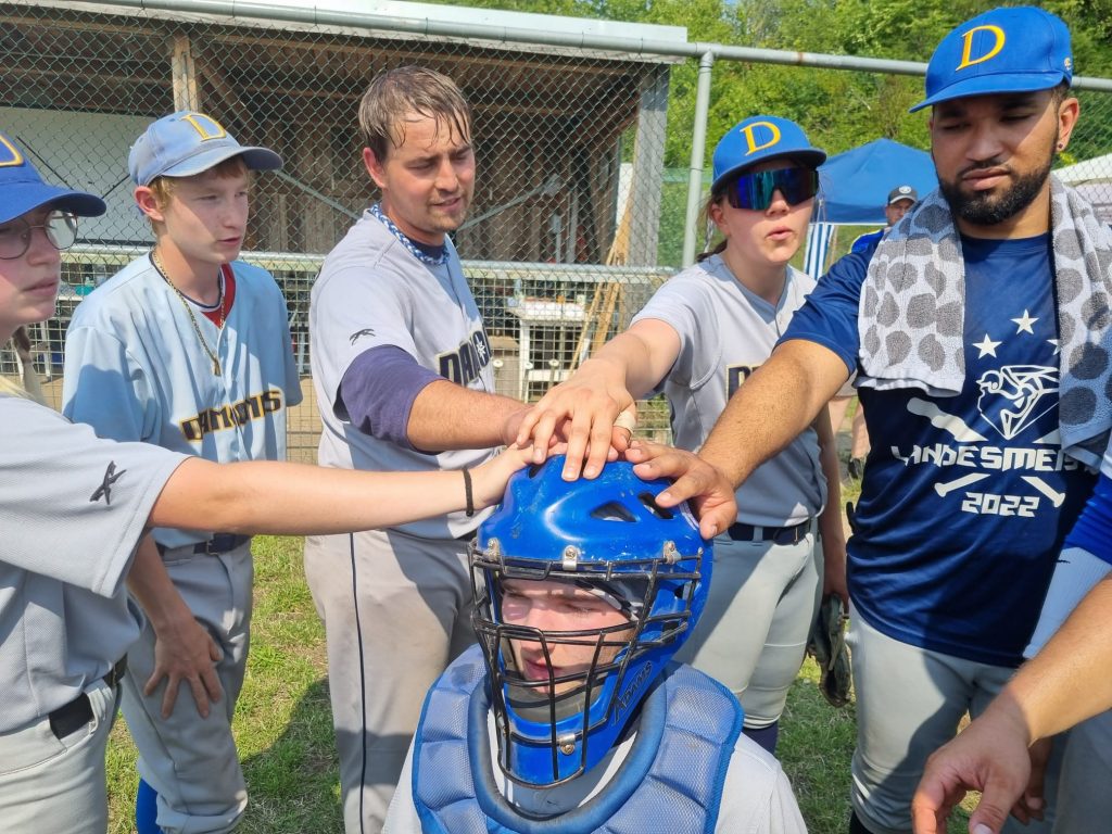 Eine Gruppe von Baseballspielern in grauen Trikots legt die Hände auf den Helm eines Mitspielers im Catcher-Outfit. Der Catcher kniet und die Teammitglieder stehen im Kreis um ihn herum.
