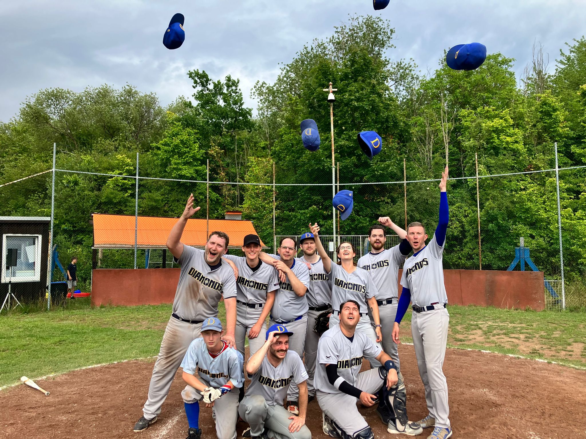 Mehrere Baseballspieler werfen freudig ihre blauen Kappen in die Luft, nachdem sie ein Spiel auf einem Baseballfeld beendet haben. Alle tragen graue Trikots und freuen sich über den Sieg.
