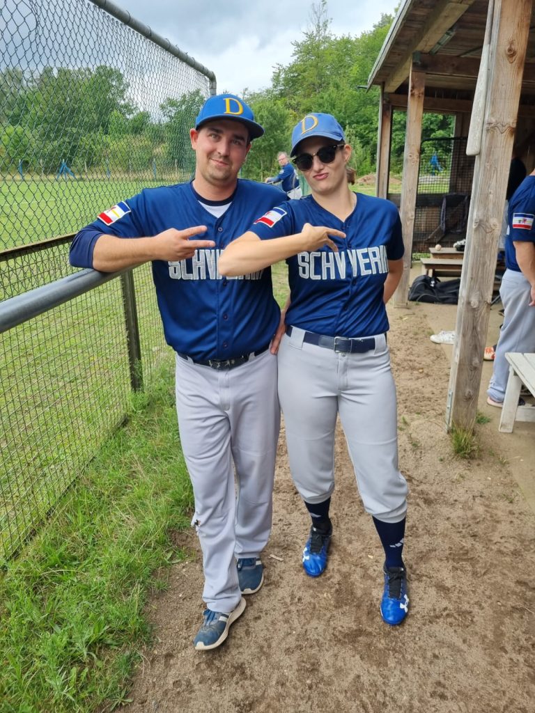 Zwei Personen in blauen Baseball-Trikots des Teams Schwerin Diamonds posieren nebeneinander und zeigen jeweils mit ihrem rechten Zeige- und Mittelfinger ein Peace-Zeichen. Neben ihnen steht ein Maschendrahtzaun, durch den man ein Spielfeld erkennen kann.