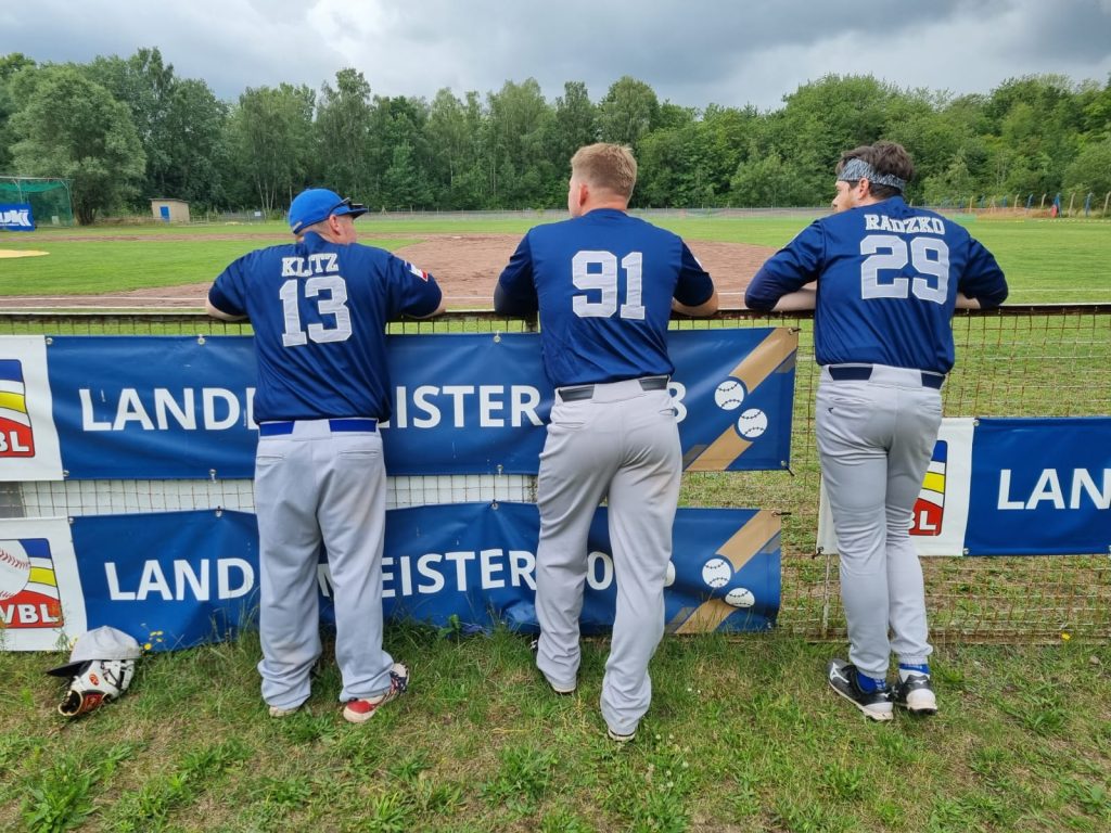 Drei Baseballspieler stehen mit dem Rücken zur Kamera, lehnen auf den Armen gestützt am Spielfeldzaun und tragen Trikots mit den Nummern 13, 91 und 29. Sie schauen auf ein Baseballfeld, vor ihnen hängt ein Banner mit der Aufschrift "Landesmeister".