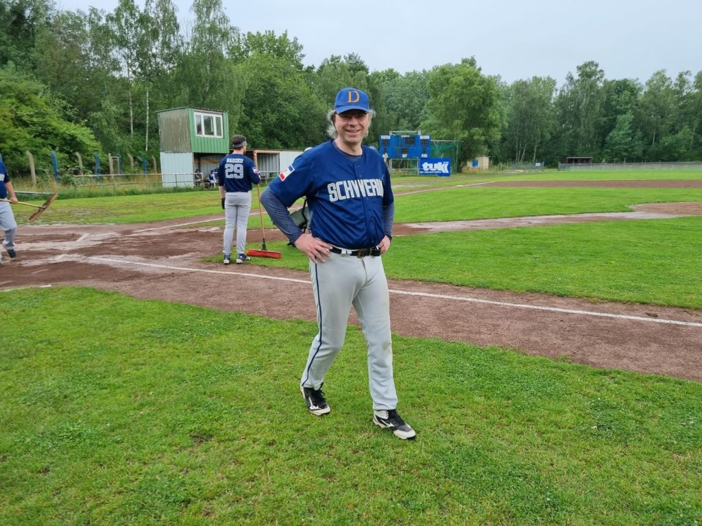 Ein Baseballspieler im blauen Schwerin Diamonds-Trikot steht auf einem Spielfeld und lächelt in die Kamera. Im Hintergrund ist ein weiteres Teammitglied sowie ein kleines Scorer-Haus sichtbar.