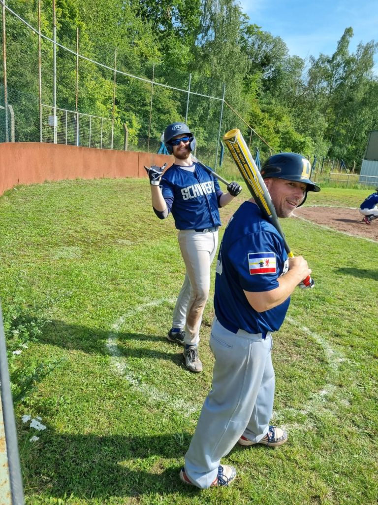 Zwei Baseballspieler des Teams Schwerin Diamonds stehen am Spielfeldrand, einer hält einen gelben Schläger auf der Schulter abgelegt, der andere hebt lächelnd die Hand. Beide tragen Helme und schauen fröhlich in die Kamera.