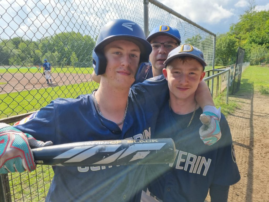 Drei Baseballspieler stehen lächelnd zusammen, einer trägt einen Helm und einen Schläger, während die anderen beiden Spieler, die neben und hinter im stehen, ein Basecap tragen. Im Hintergrund befindet sich ein Spielfeld mit weiteren Spielern.