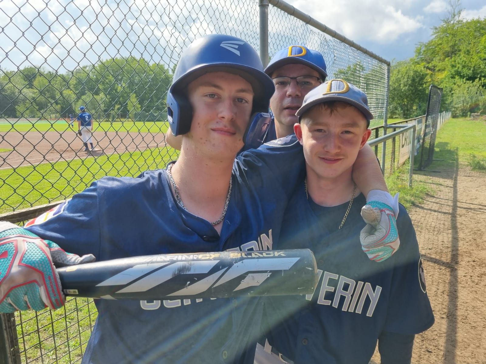 Drei Baseballspieler stehen lächelnd zusammen, einer trägt einen Helm und einen Schläger, während die anderen beiden Spieler, die neben und hinter im stehen, ein Basecap tragen. Im Hintergrund befindet sich ein Spielfeld mit weiteren Spielern.