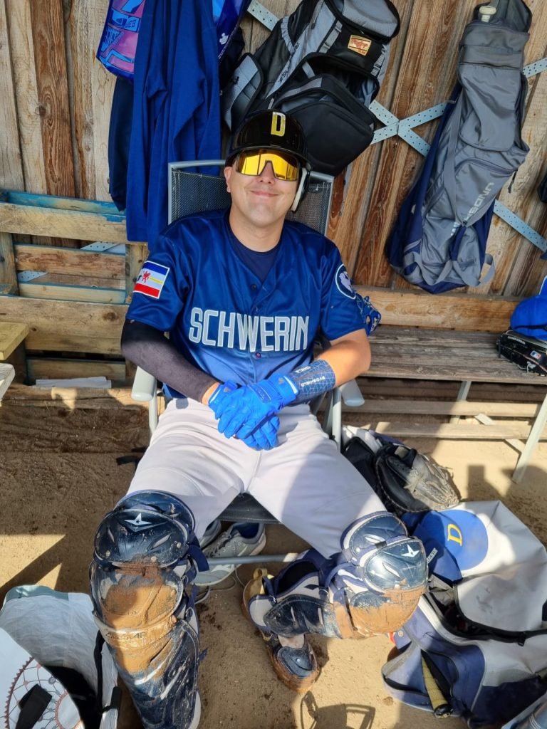 Ein Baseball-Catcher sitzt in der Teamzone auf einem Stuhl, trägt komplette Schutzausrüstung und ein Schwerin-Trikot. Hinter ihm hängen Taschen und Ausrüstungsgegenstände an einer Holzwand.