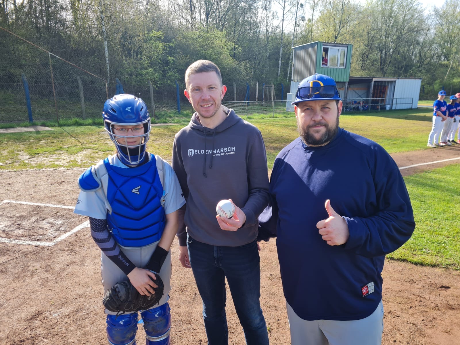 Drei Personen stehen auf einem Baseballfeld: Links ein Catcher in kompletter Schutzausrüstung, in der Mitte eine Person mit einem Baseball in der Hand und rechts ein Trainer, der den Daumen hochhält. Im Hintergrund sind weitere Spieler sowie eine grüne Landschaft mit Bäumen und einer Hütte zu sehen.